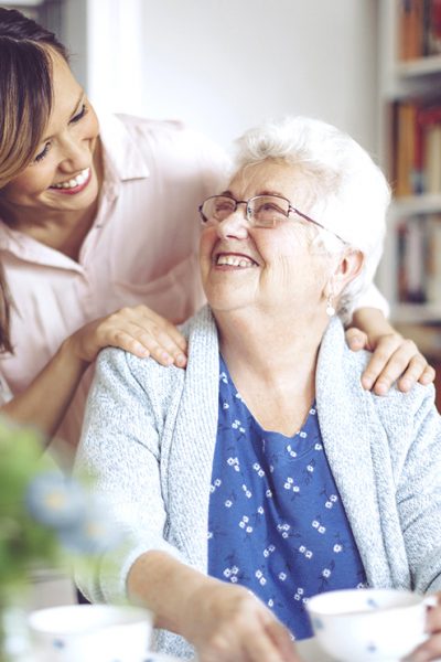 Social worker is visiting a senior woman in her own apartment. They are celebrating the elderly lady's birthday. The kind nurse is hugging the senior woman over her shoulder.